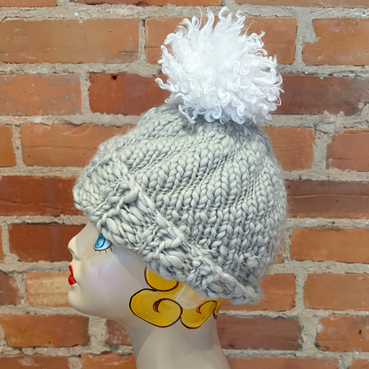 A light gray chunky knit beanie with a white curly pom-pom, displayed on a decorative mannequin head against a red brick wall background.