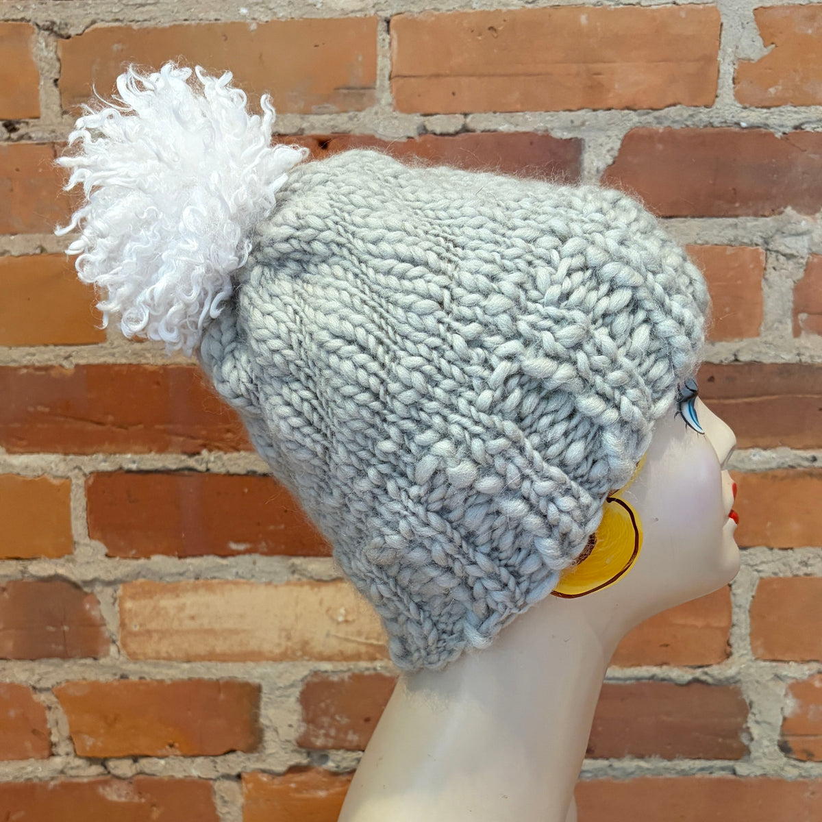 A chunky knit beanie hat in a light heather gray color with a large fluffy white pom-pom, displayed on a mannequin head against a brick wall background.