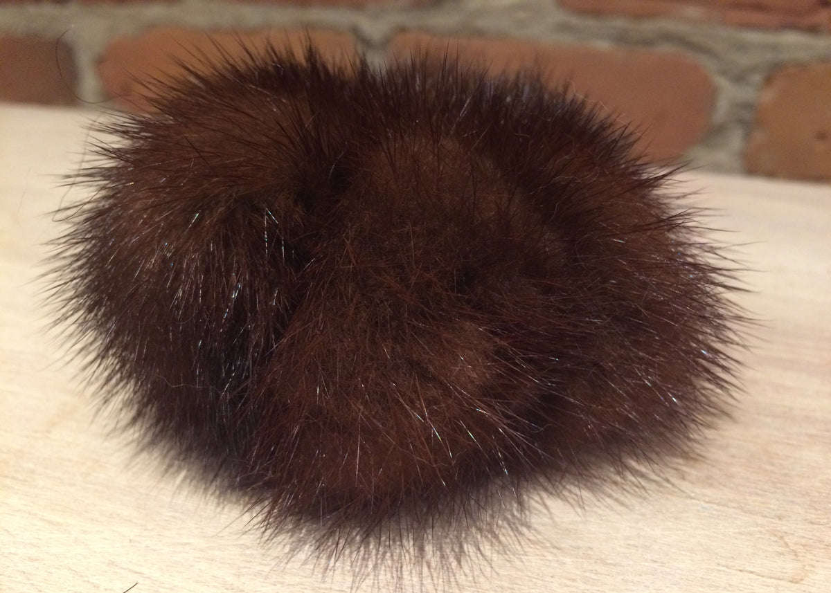 A dark brown fluffy mink fur pom-pom accessory resting on a light wood surface with a blurred brick wall background.