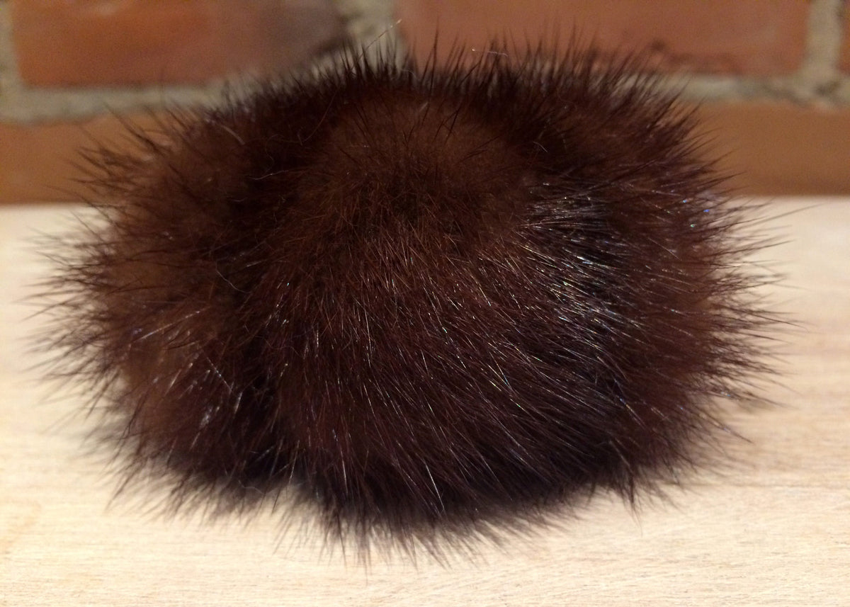 A dark brown fluffy mink fur pom-pom accessory resting on a light wood surface with a brick wall background.