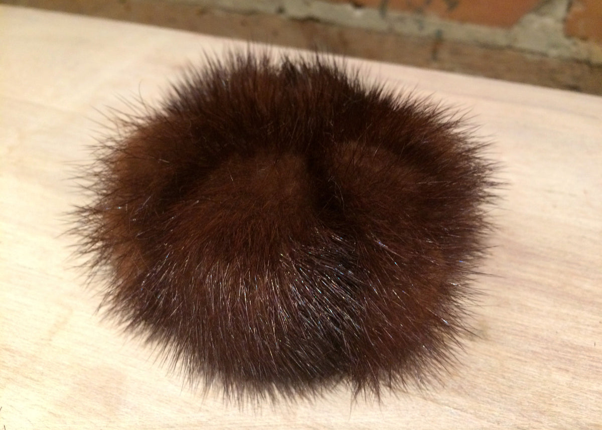 A dark brown fluffy mink fur pom-pom accessory resting on a light-colored wooden surface.