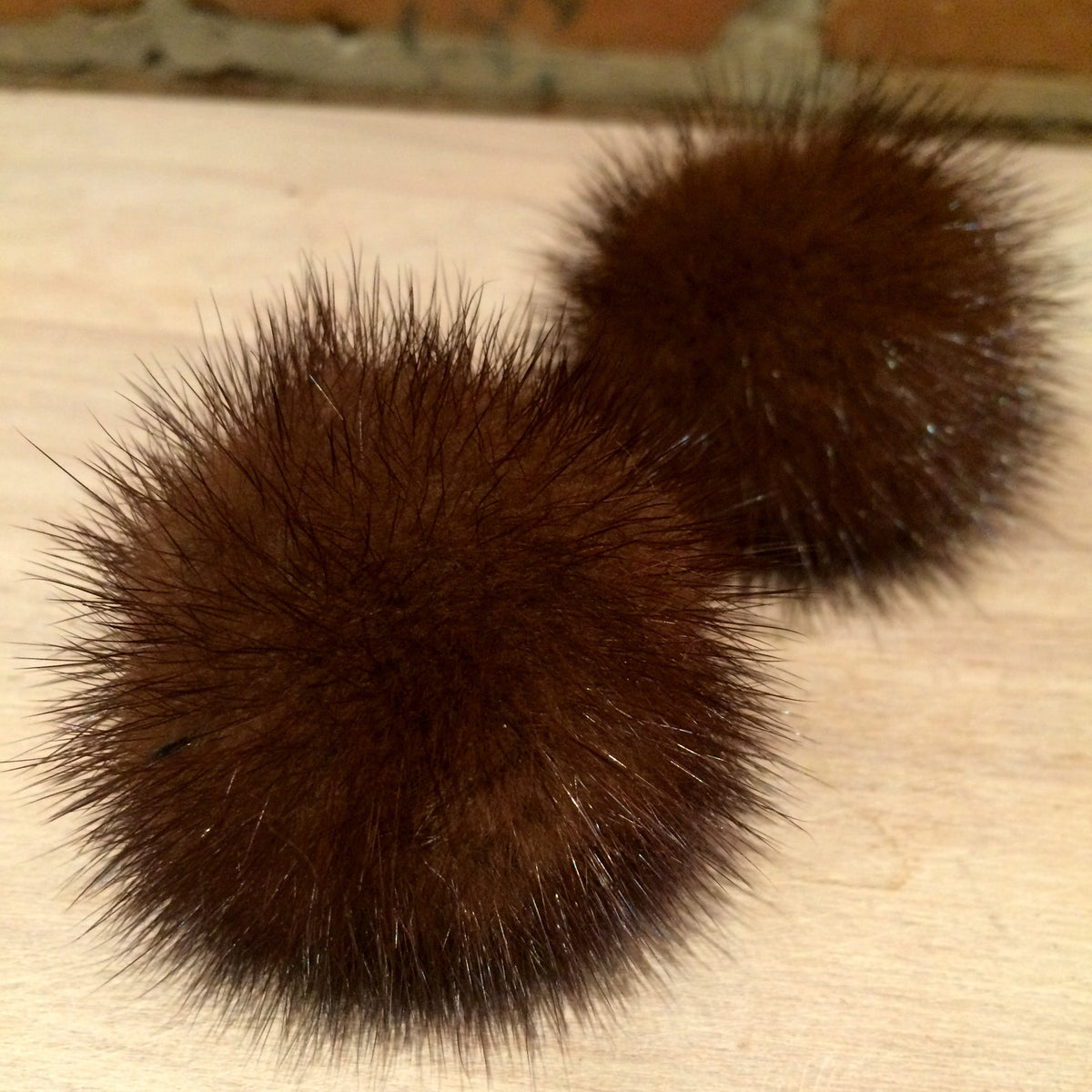 Two dark brown furry pom-poms on a light wood surface with a brick wall in the background.