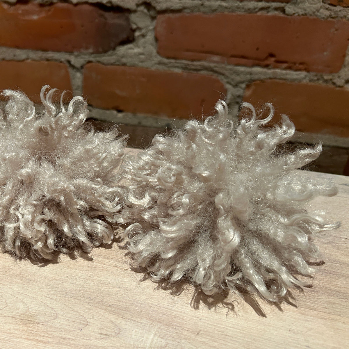 Two fluffy curly faux fur pom-poms in a light oatmeal beige color, resting on a light wood surface with a rustic brick wall background.