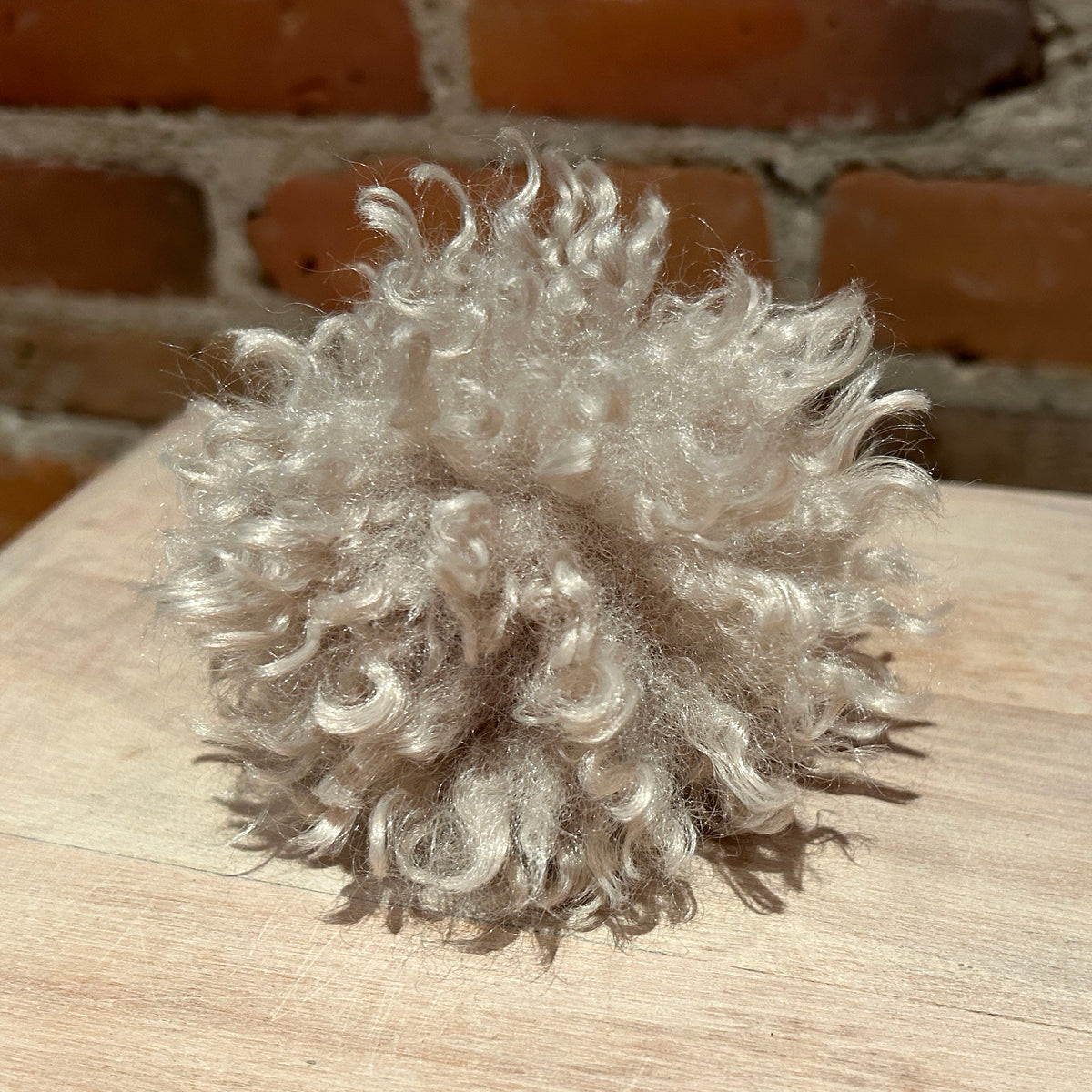 A fluffy, curly pom-pom made from light silver-gray synthetic fibers, resting on a light wood surface with a blurred brick wall background.