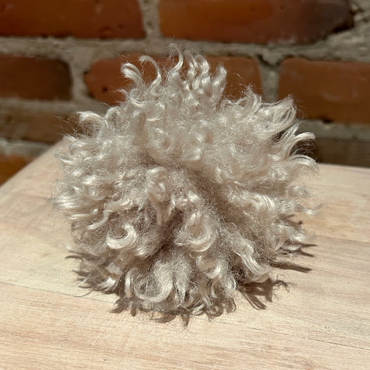 A fluffy, curly pom-pom made from light silver-gray synthetic fibers, resting on a light wood surface with a blurred brick wall background.