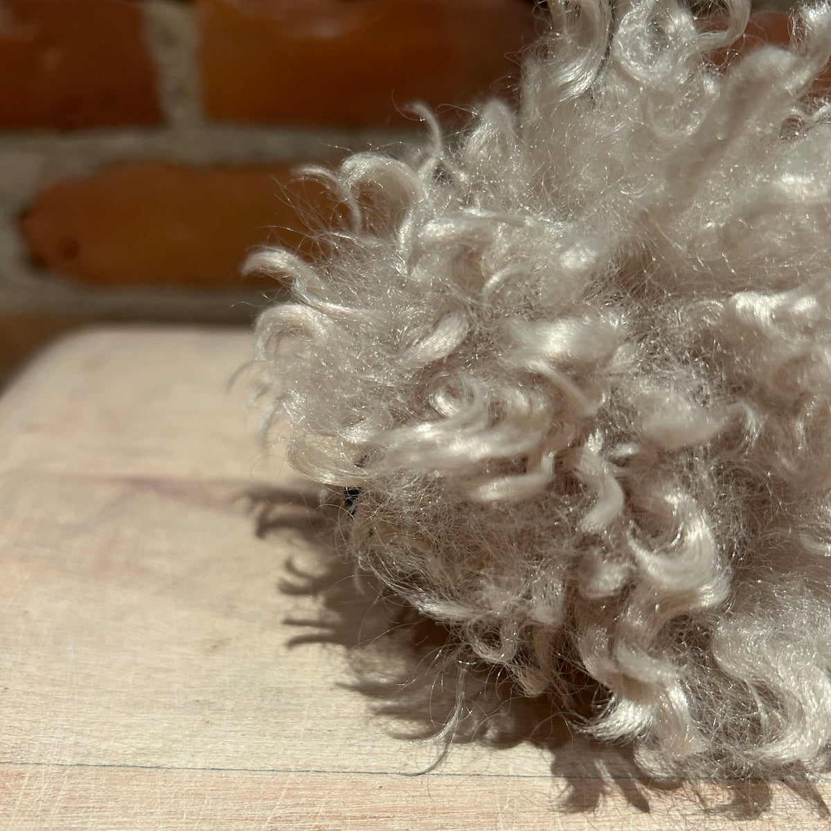 A close-up of curly, fluffy light silver-gray wool fibers resting on a light wood surface with a blurred brick wall in the background.