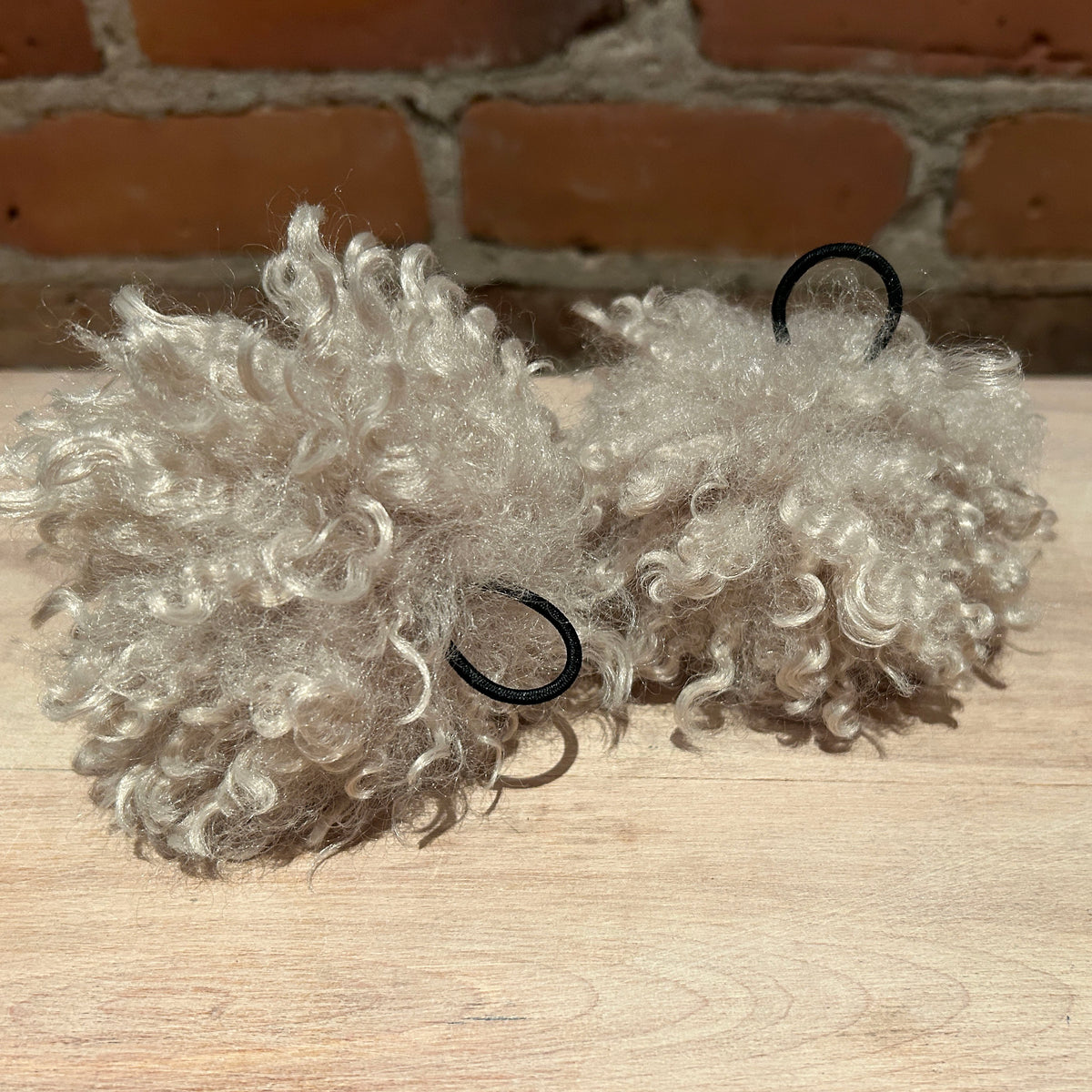 Two fluffy curly faux fur pom poms in a light ash-blonde color, each attached to a black elastic hair tie, displayed on a light wood surface with a brick wall background.
