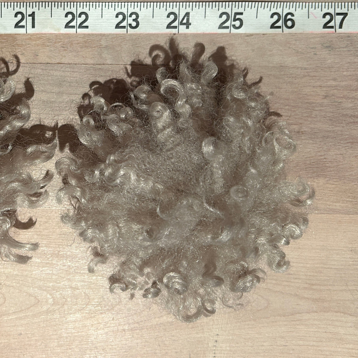 A pile of curly, light brown to gray faux fur pom poms on a light wood surface, with a measuring ruler visible at the top for scale.