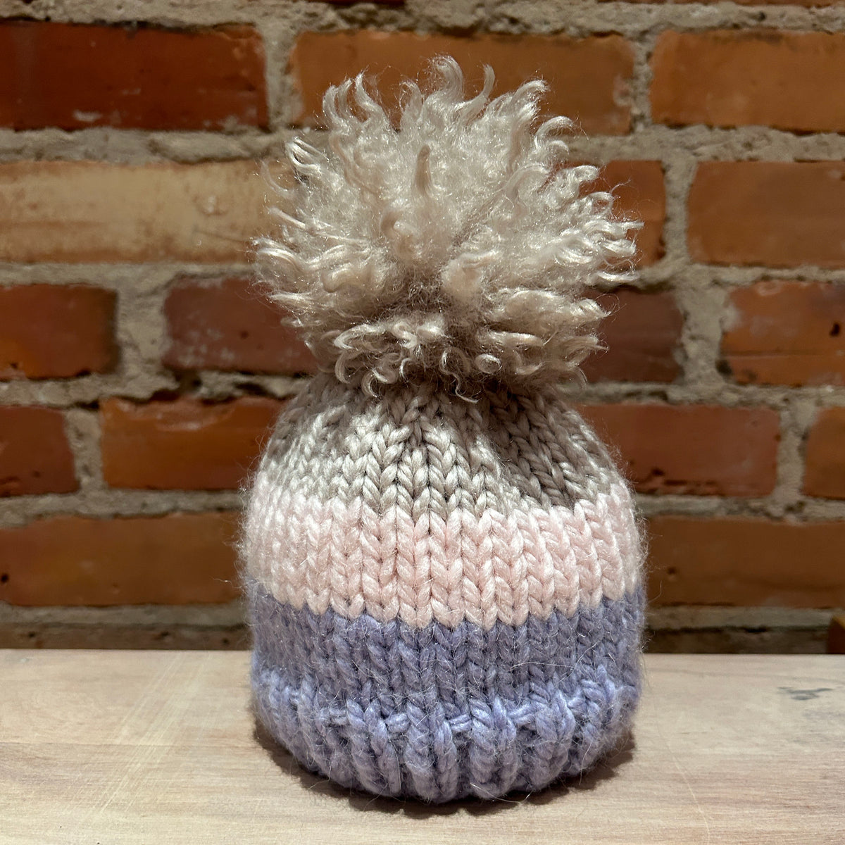 A hand-knit pom-pom beanie hat with horizontal stripes of light gray, pale pink, and dusty lavender, topped with a fluffy cream pom-pom, set against a rustic brick wall background.
