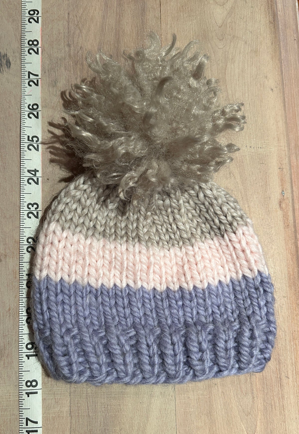 A hand-knit beanie hat with three horizontal stripes in taupe, pale pink, and dusty lavender, topped with a large fluffy taupe pom-pom. The beanie is laid flat on a light wood floor next to a measuring tape ruler.