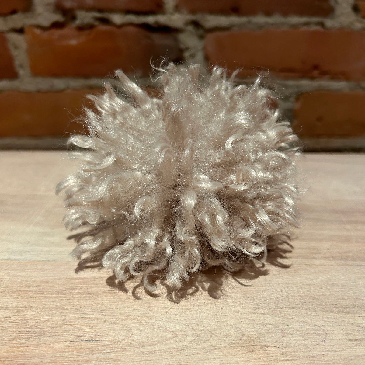A fluffy, curly pom-pom accessory made from light beige faux fur or wool, resting on a light wood surface with a brick wall in the background.