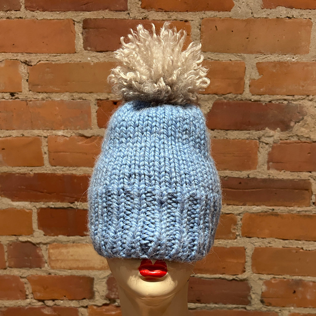 A light blue hand-knit beanie with a fluffy off-white pom-pom, displayed on a mannequin head with red lips in front of a red brick wall.