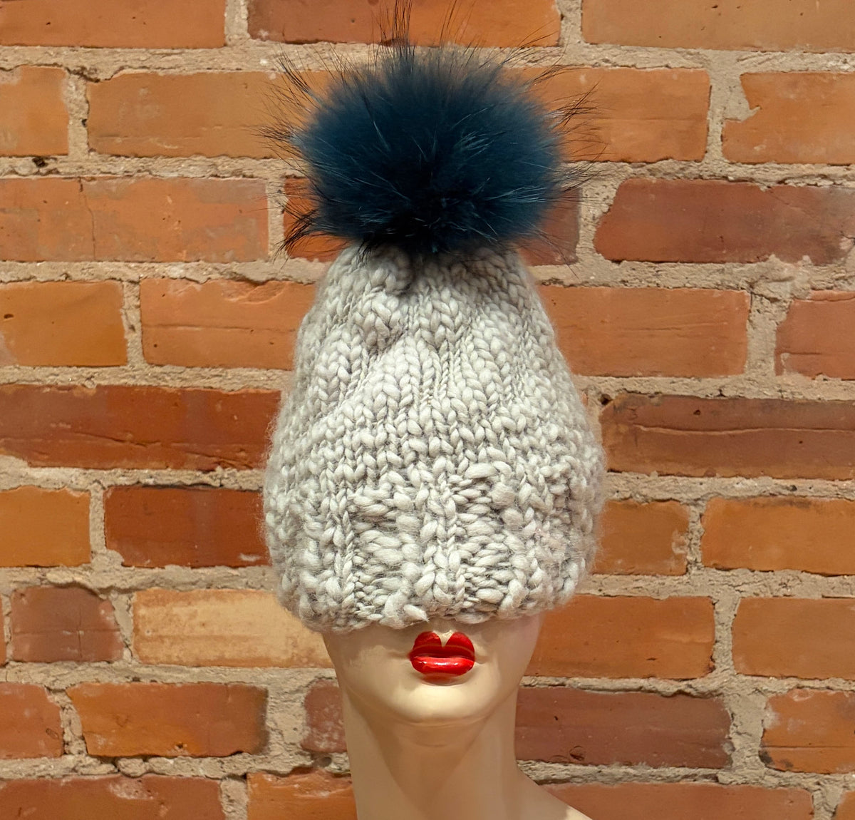 A chunky knit beanie hat in off-white with a large dark teal finnish raccoon fur pom-pom, displayed on a mannequin head with red lips against a brick wall background.