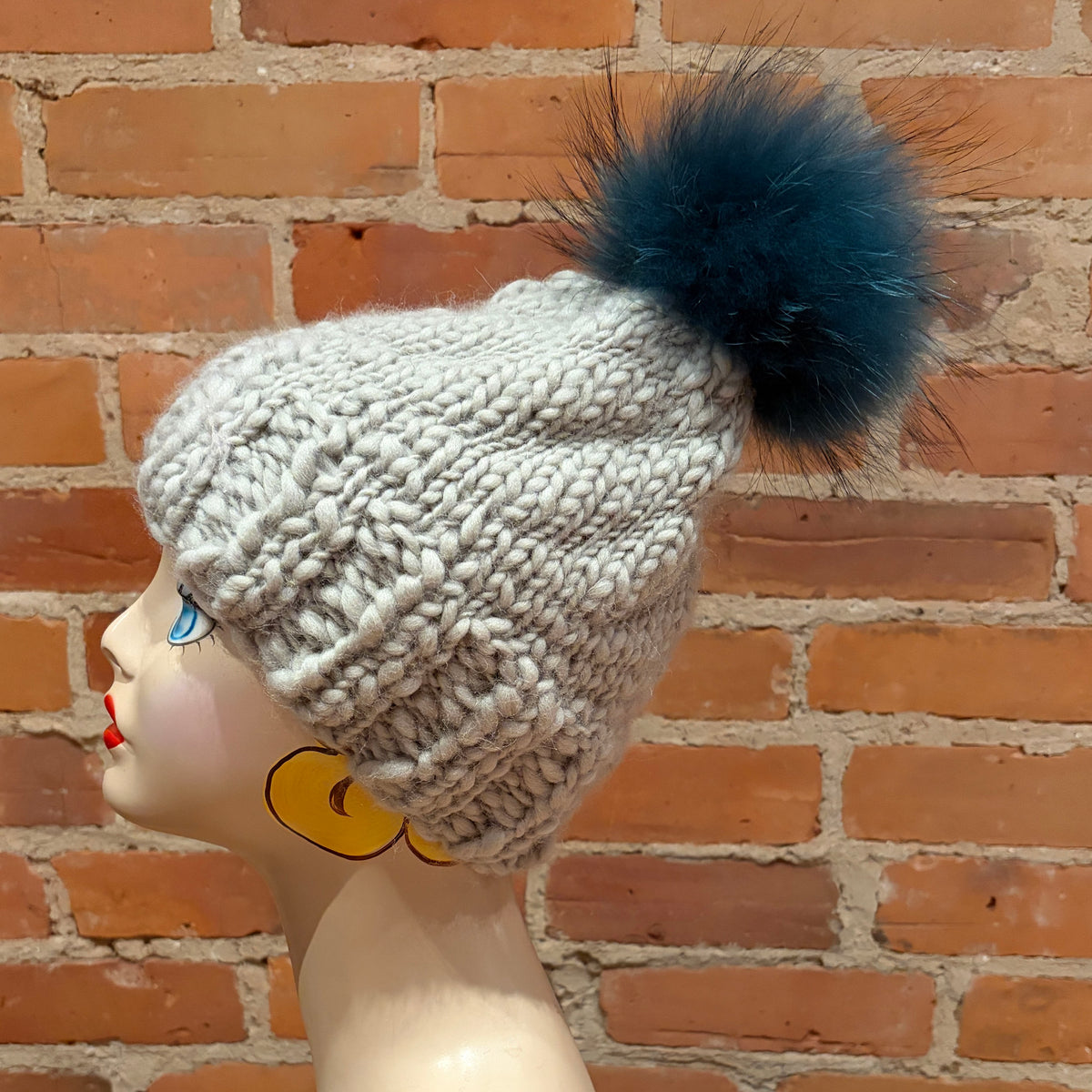 A chunky knit beanie hat in light gray with a large dark teal finnish raccoon fur pom-pom, displayed on a mannequin head against a brick wall background.