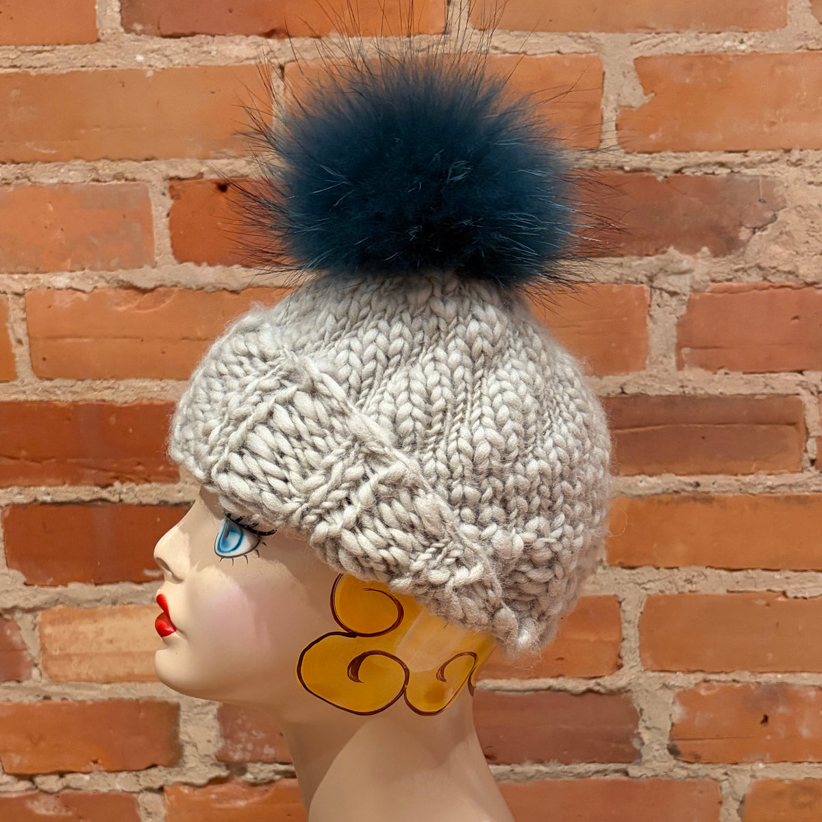 A chunky knit winter hat in light heather gray with a large dark teal finnish raccoon fur pom-pom, displayed on a mannequin head against a red brick wall background.