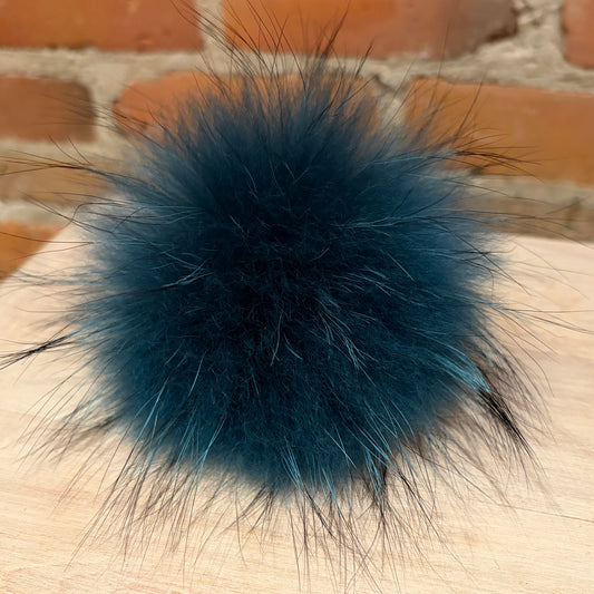A fluffy dark teal finnish raccoon fur pom-pom with black and blue-gray tips, resting on a light wood surface with a blurred red brick wall background.
