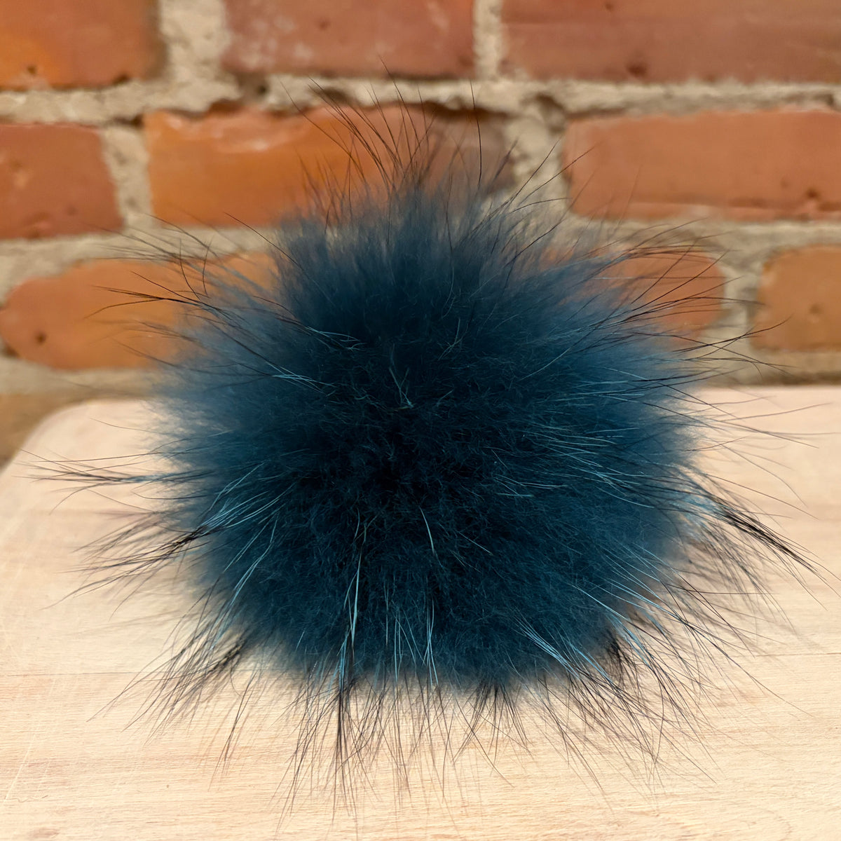 A fluffy finnish raccoon-fur pom pom in a deep teal blue color, resting on a light wood surface with a brick wall background.