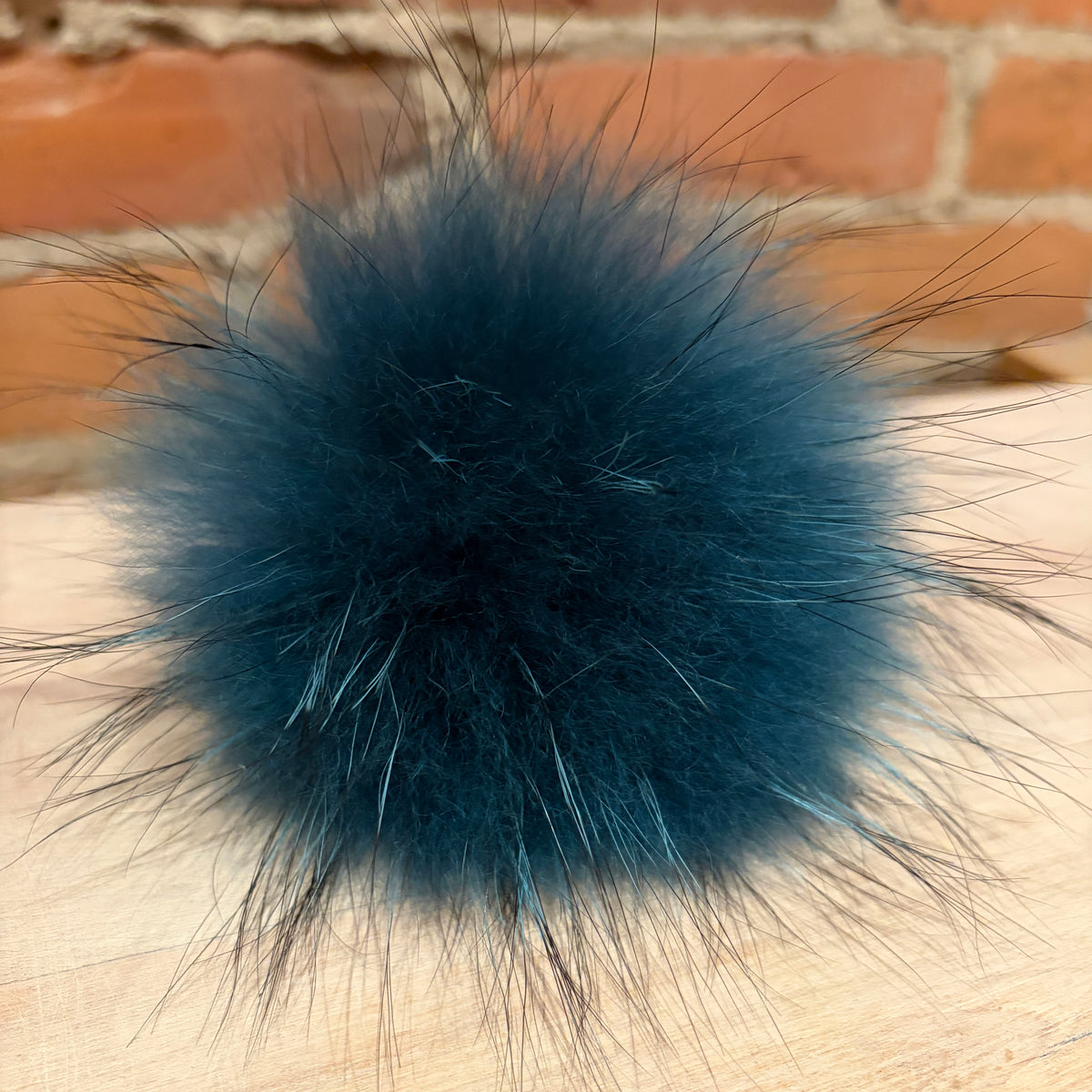 A fluffy dark teal finnish raccoon fur pom-pom accessory resting on a light wood surface with a blurred brick wall background.