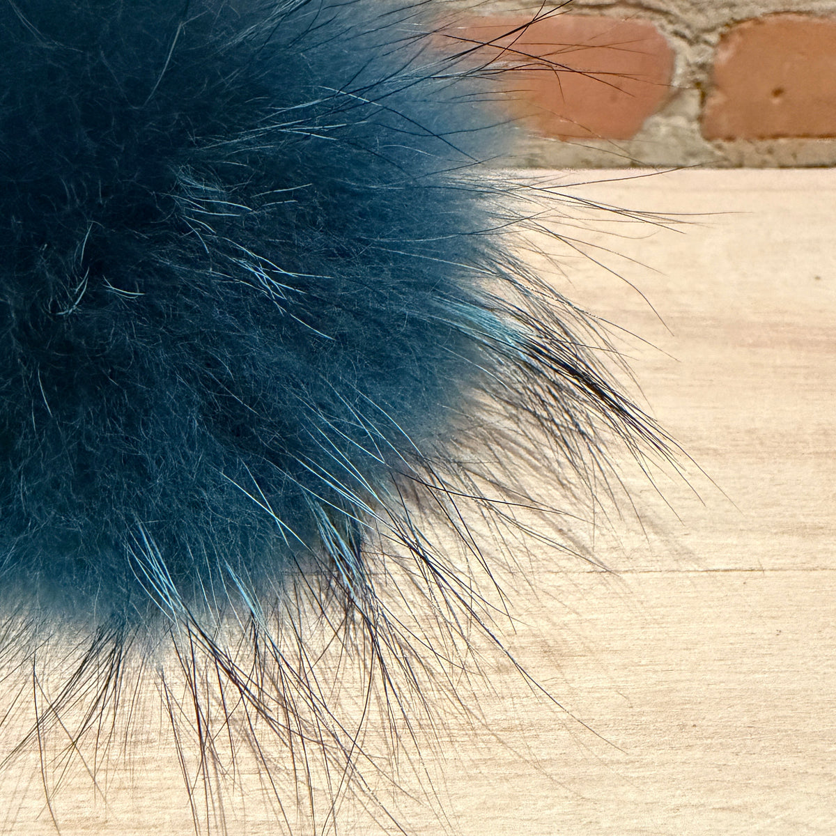 A close-up of a fluffy dark blue finnish raccoon fur pom-pom resting on a light wood surface with a blurred brick wall in the background.