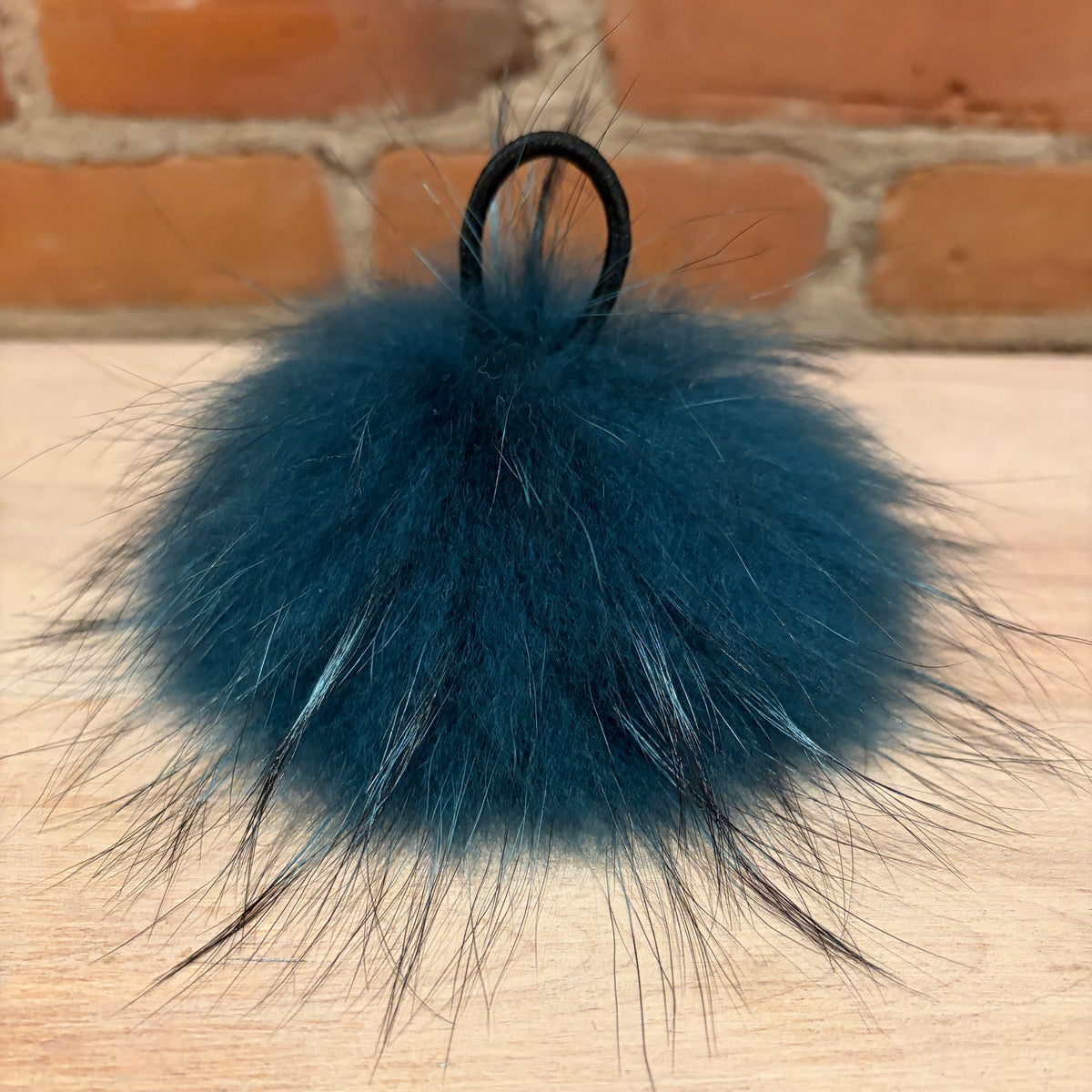 A dark teal blue fluffy finnish raccoon fur pom-pom keychain with a black elastic loop, resting on a light wood surface with a brick wall background.