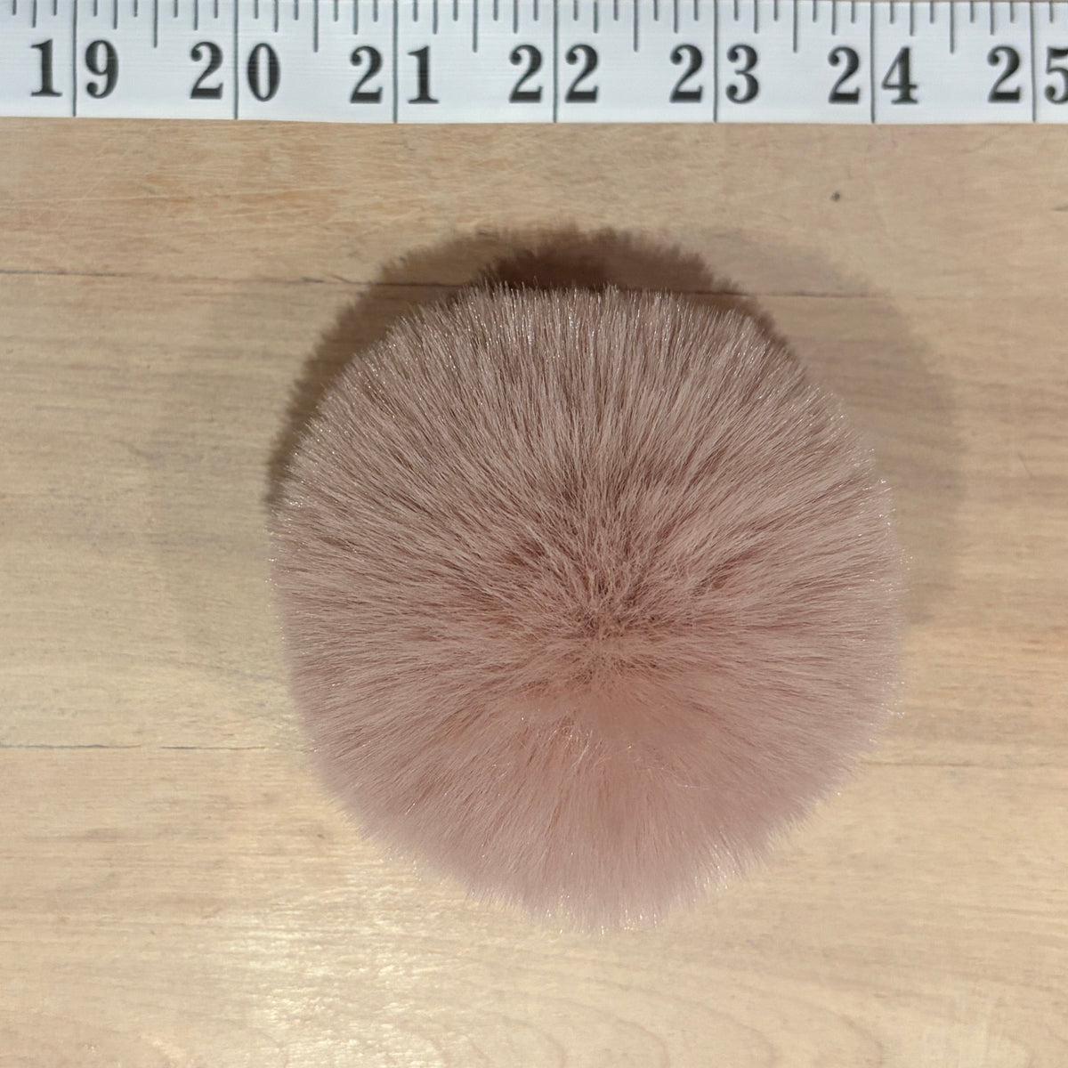 A round, fluffy faux fur pom-pom in a dusty pink color, shown on a light wood surface with a measuring tape ruler at the top for scale.