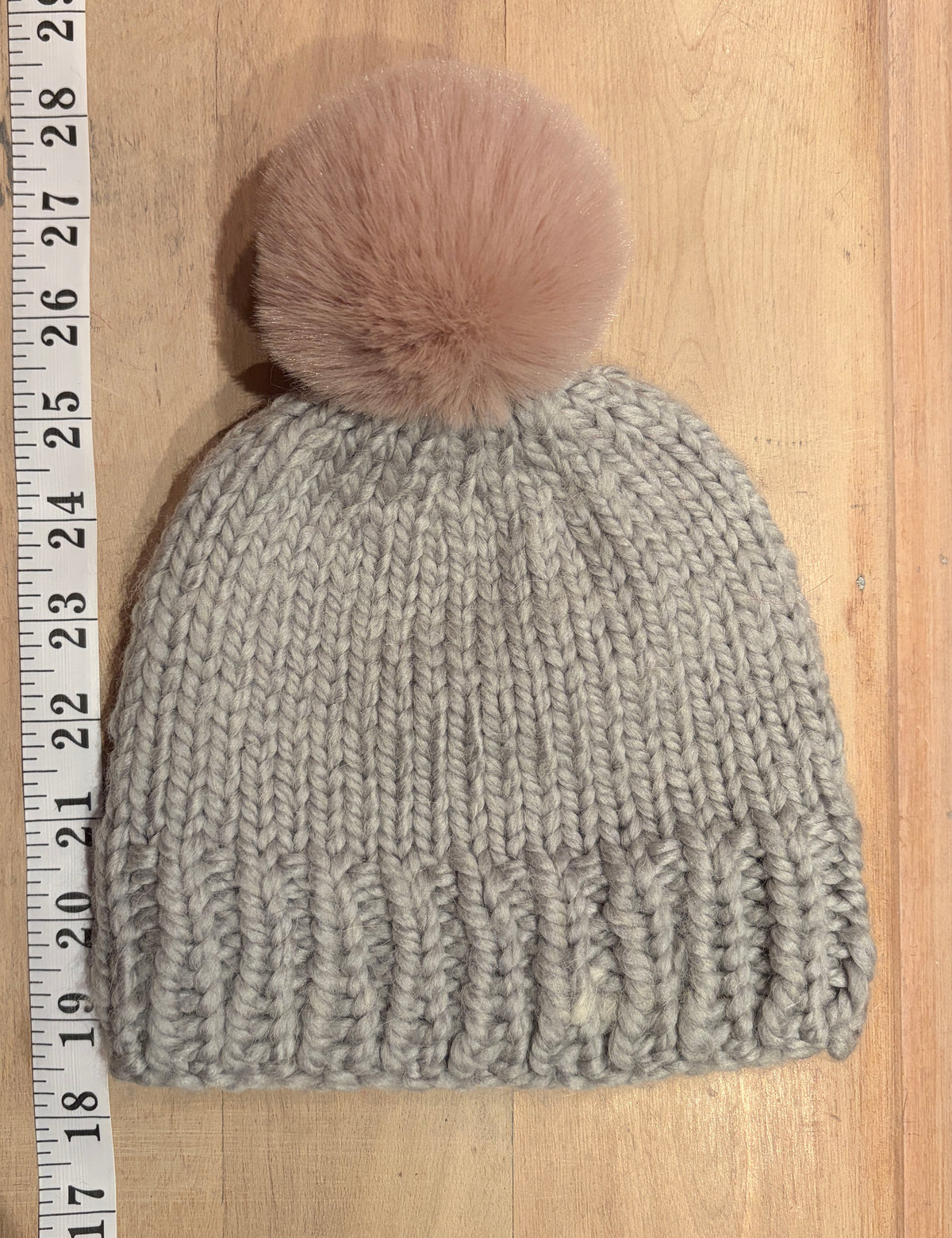 A light gray ribbed knit beanie with a dusty pink faux fur pom-pom, shown on a light wood background next to a measuring tape ruler.
