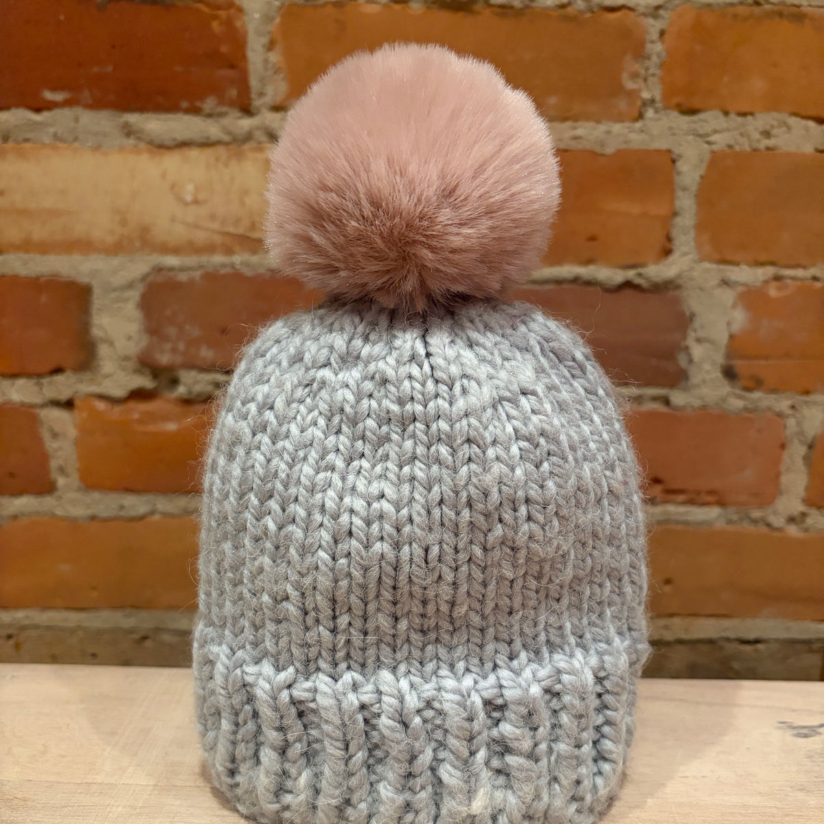 A light gray chunky knit beanie hat with a large dusty pink faux fur pom-pom, displayed on a wooden surface against a rustic brick wall background.
