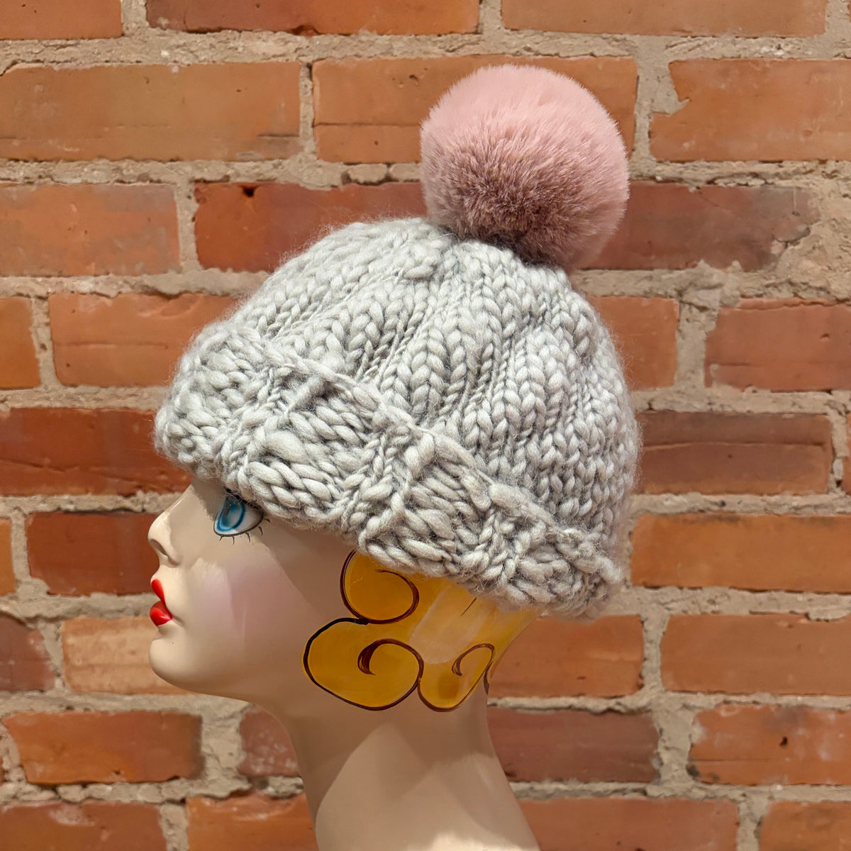 A chunky knit winter hat in light gray with a soft pink faux-fur pom-pom, displayed on a mannequin head against a red brick wall background.