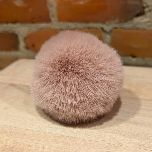 A fluffy pom-pom accessory in a dusty pink color, resting on a light wood surface with a rustic brick wall in the background.
