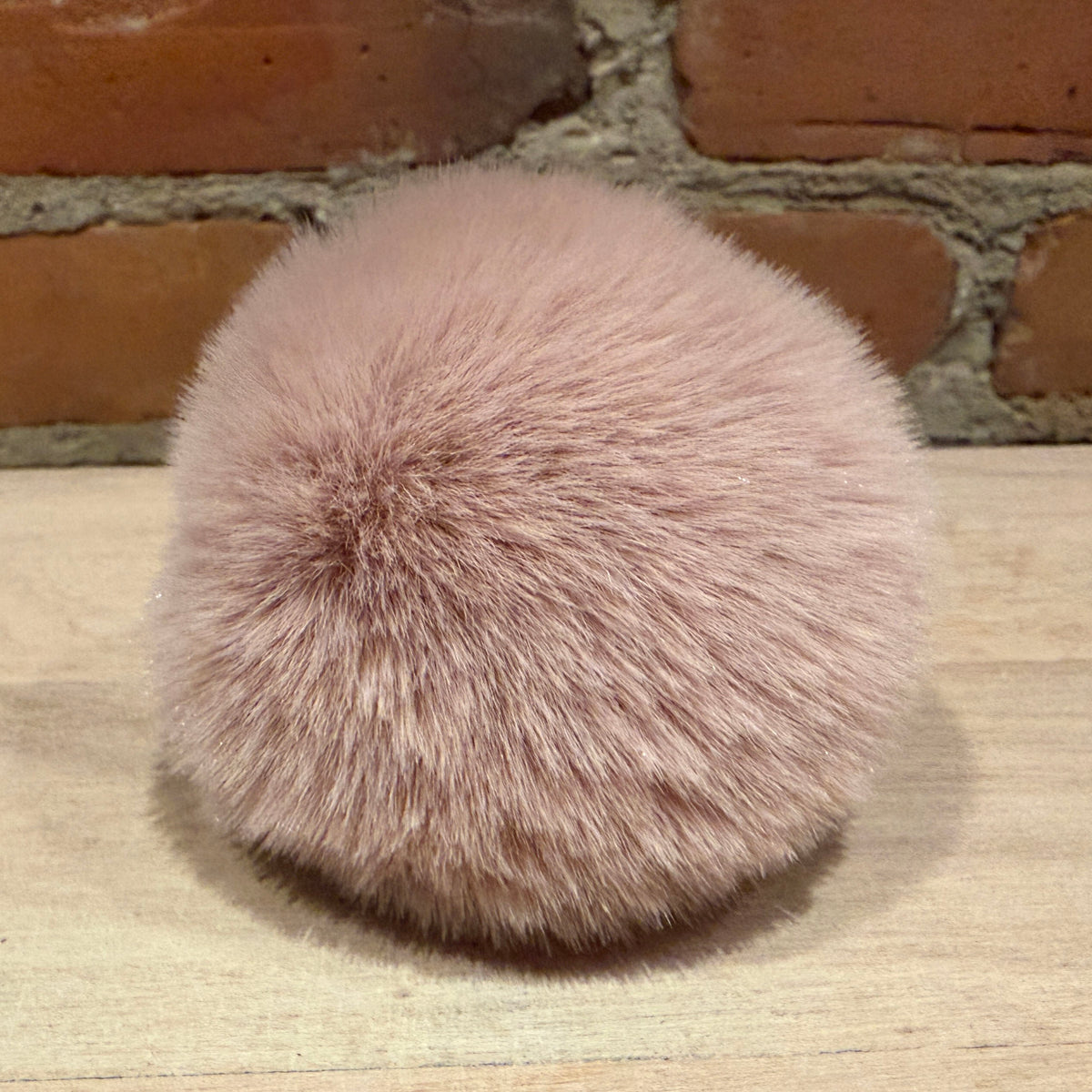 A fluffy pom-pom accessory in a dusty pink color, resting on a light wood surface with a rustic brick wall background.