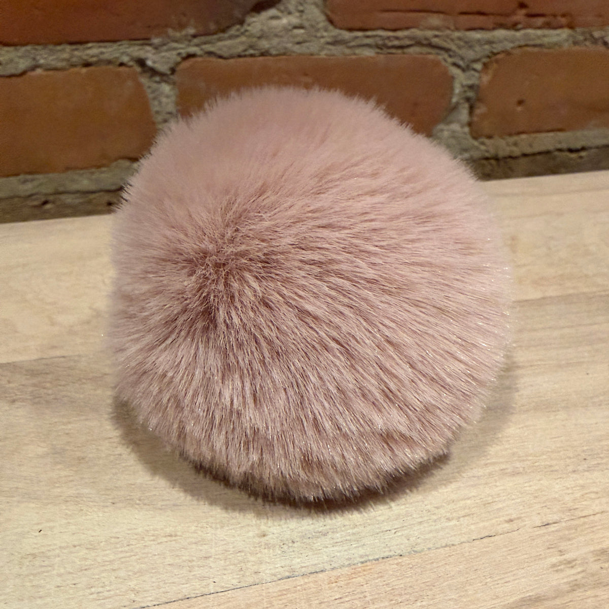 A round, fluffy faux fur pom-pom in a dusty pink color, resting on a light-colored wooden surface with a brick wall background.