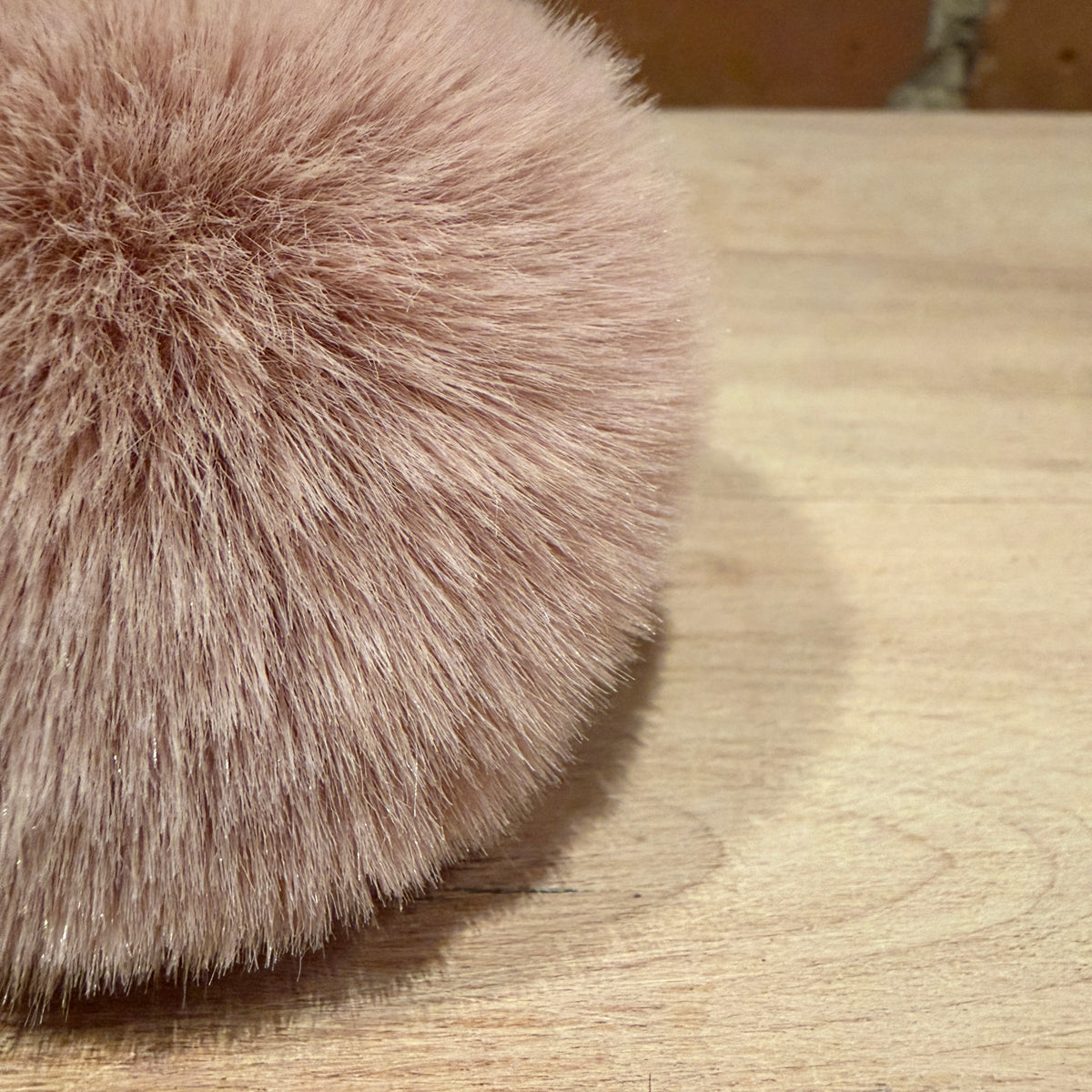 A close-up of a fluffy faux fur pom-pom in a dusty pink color, resting on a light wood surface.