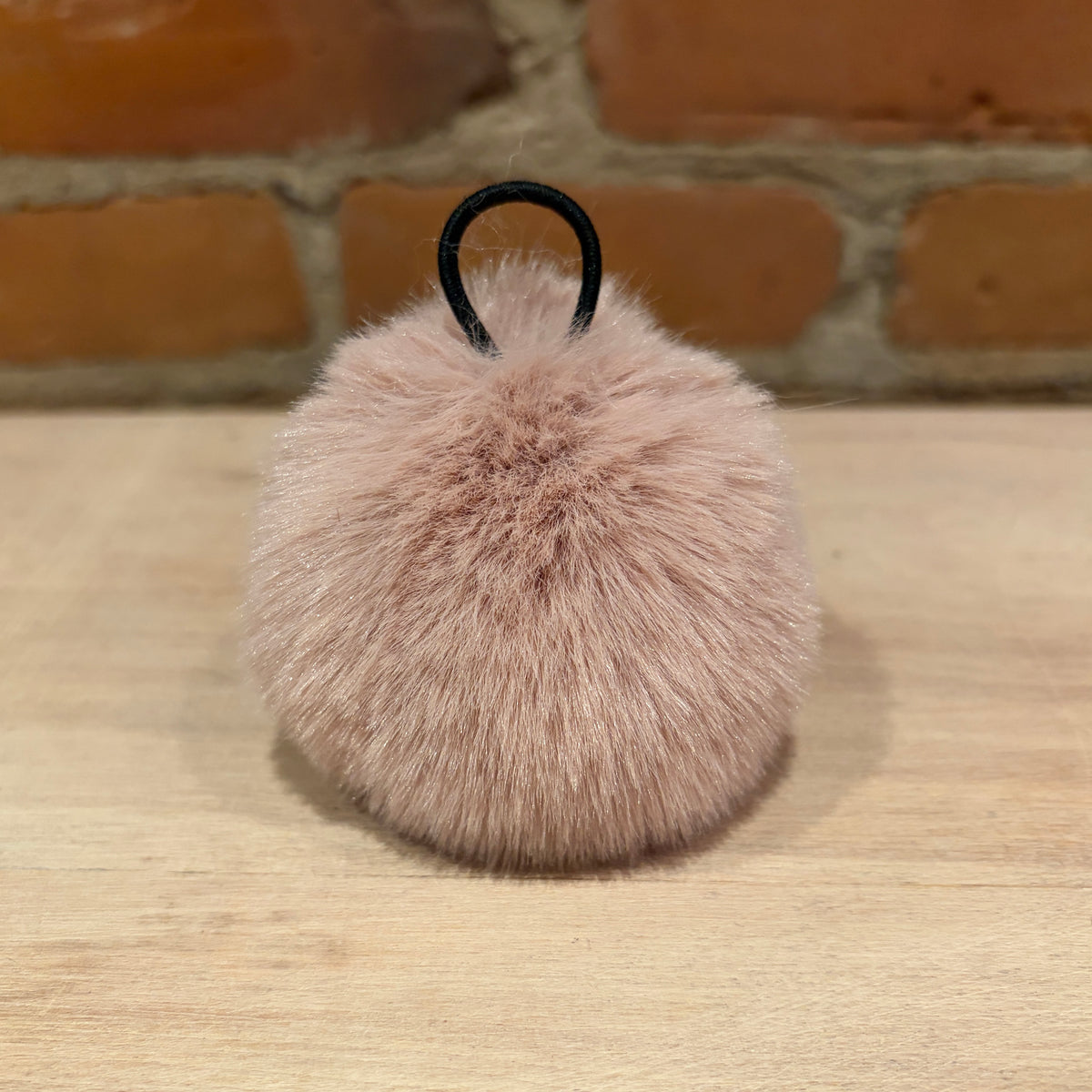 A fluffy pom-pom keychain in a dusty pink color with a black plastic loop, resting on a light wood surface with a brick wall background.