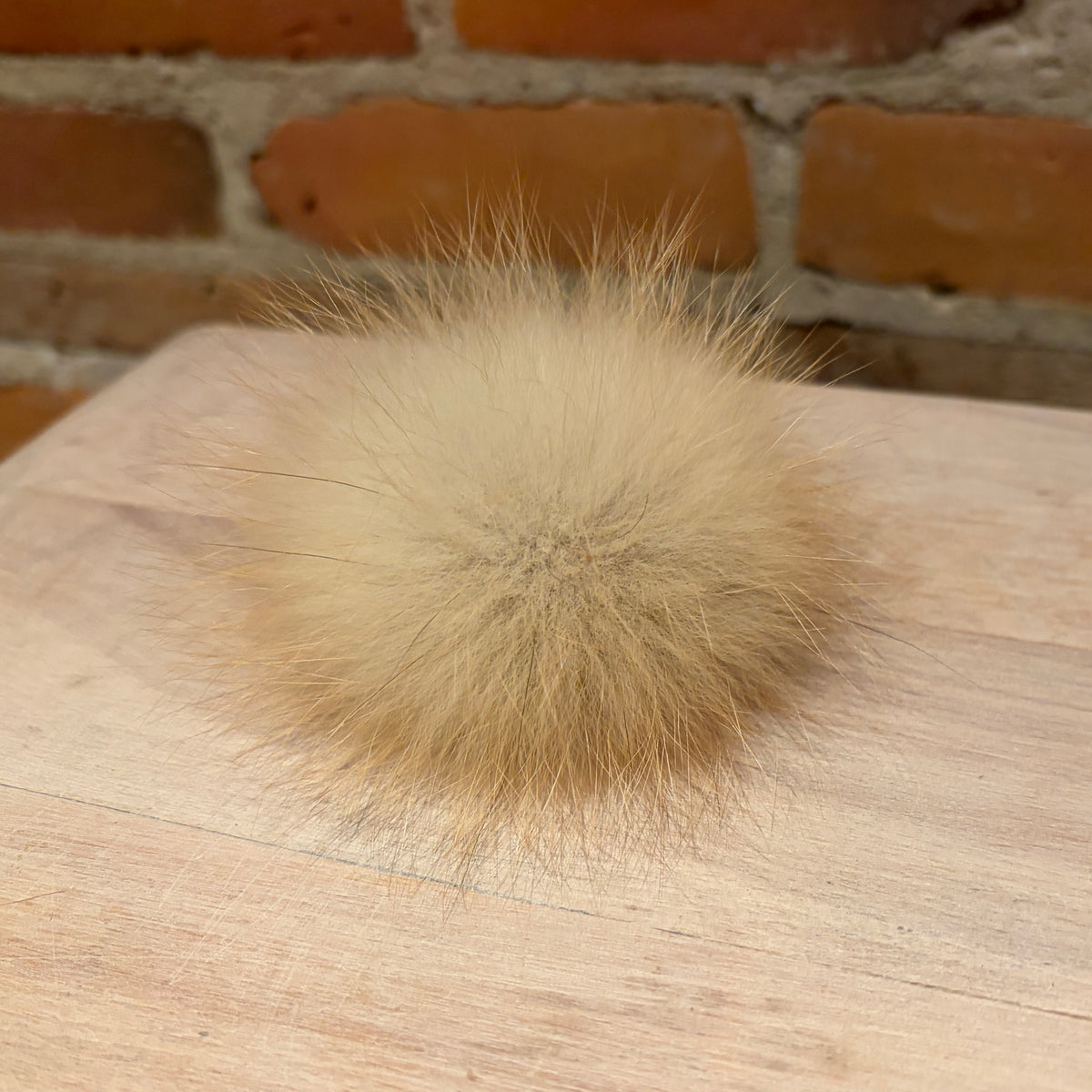 A fluffy, round fox -fur pom-pom ball in a light tan/beige color, resting on a light wood surface with a blurred brick wall background.
