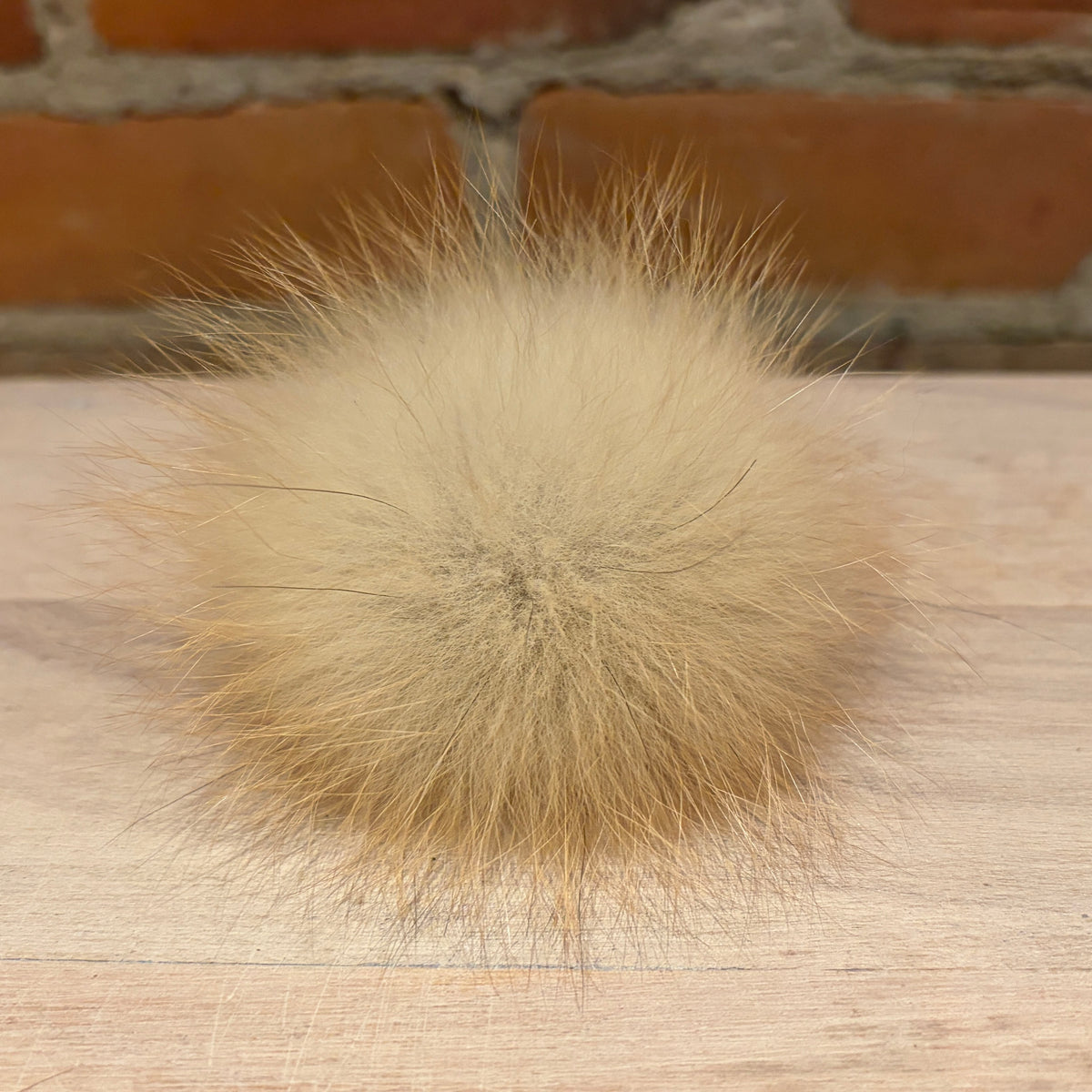 A fluffy beige fox fur pom-pom ball resting on a light wood surface with a blurred brick wall background.