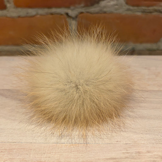 A fluffy beige fox fur pom-pom ball resting on a light wood surface with a blurred brick wall background.