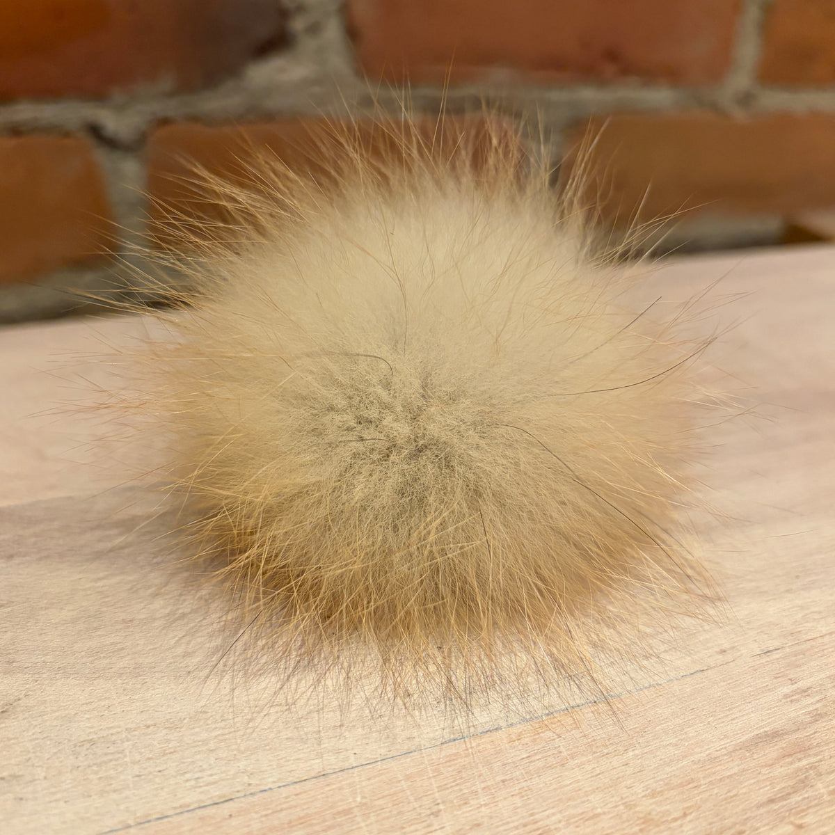 A fluffy fox -fur pom-pom ball in a light beige color, resting on a light wood surface with a blurred brick wall background.