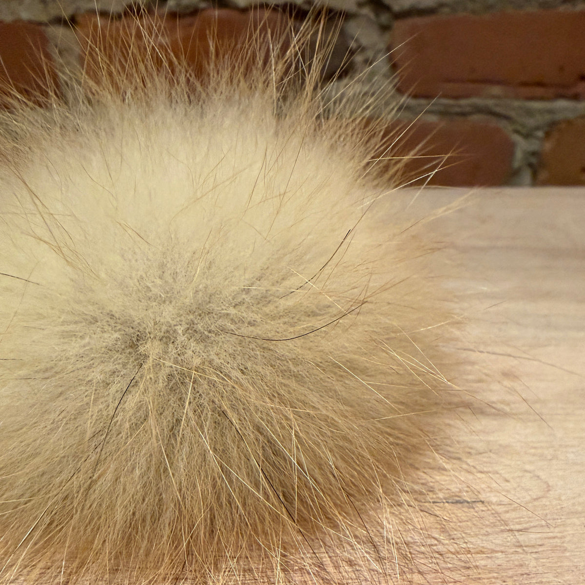 A close-up of a fluffy pom-pom accessory in a light beige or cream color, resting on a light wood surface with a blurred brick wall background.
