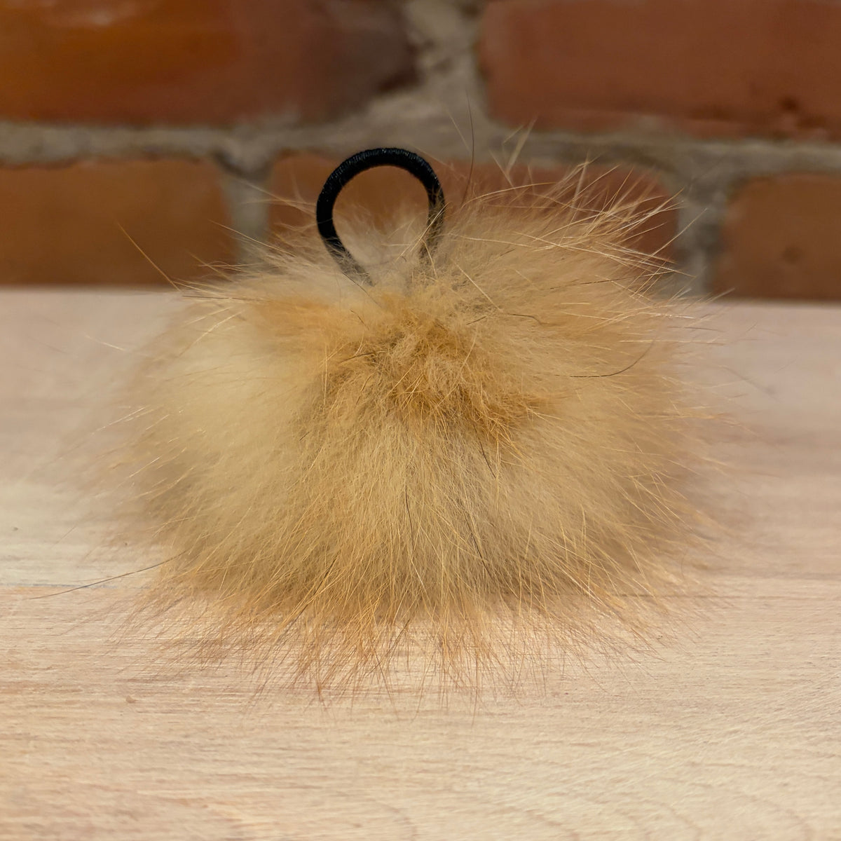 A fluffy fox -fur pom-pom keychain or bag charm in a warm tan and light brown shade, featuring a black plastic ring at the top, resting on a light wood surface with a blurred brick wall background.