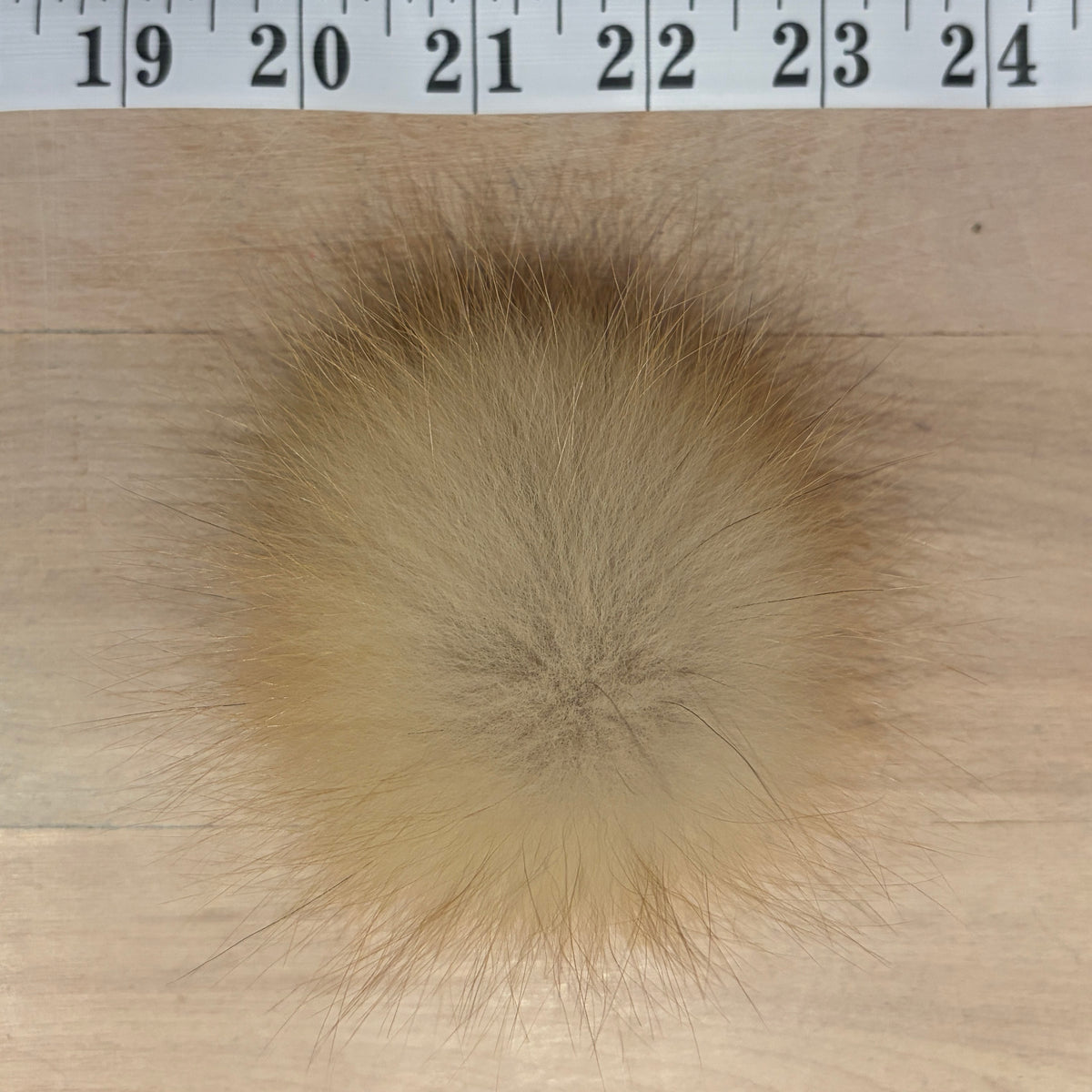 A round beige fox fur pom-pom with darker brown tips, shown on a light wood surface with a measuring tape ruler at the top for scale.