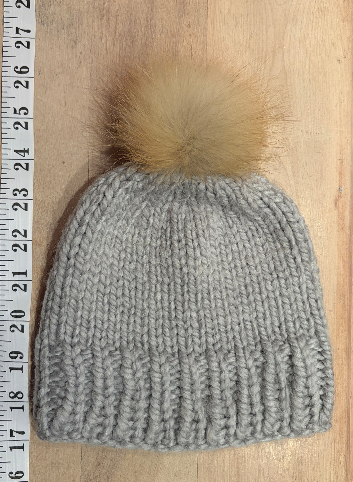 A light gray chunky knit beanie with a small tan fox fur pom-pom, laid flat on a light wood surface next to a measuring tape ruler.