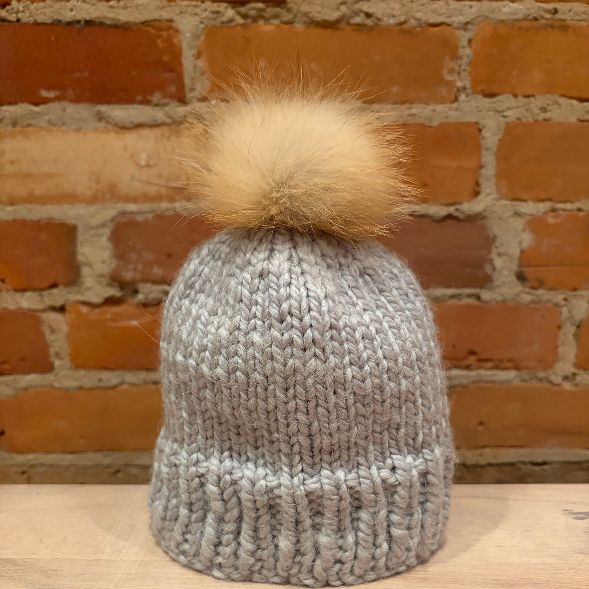 A chunky knit gray beanie with a small  fluffy beige fox fur pom-pom, displayed on a wooden surface against a rustic red brick wall background.