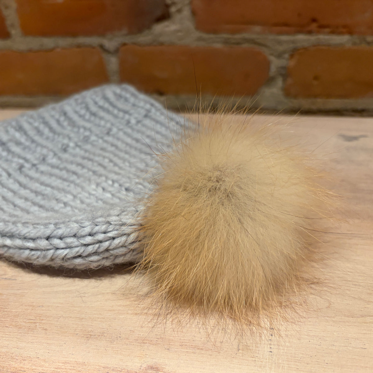 A close-up of a light gray knit beanie with a fluffy tan fox -fur pom-pom, resting on a light wood surface with a brick wall in the background.
