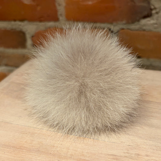 A fluffy pom-pom ball in a light taupe-gray color, resting on a light wood surface with a blurred brick wall background.