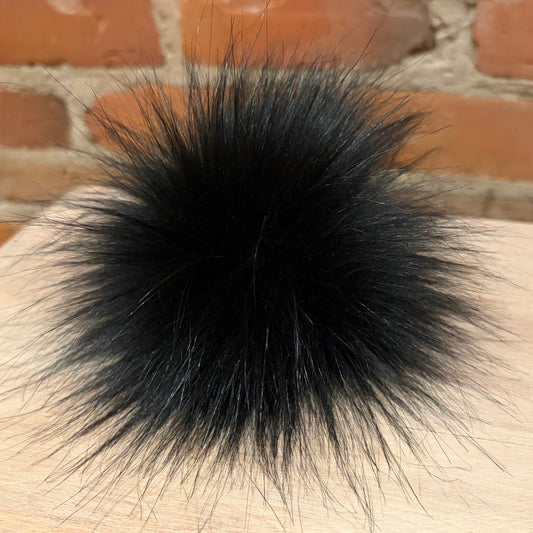 A fluffy black faux fur pom-pom accessory resting on a light wood surface with a blurred brick wall background.