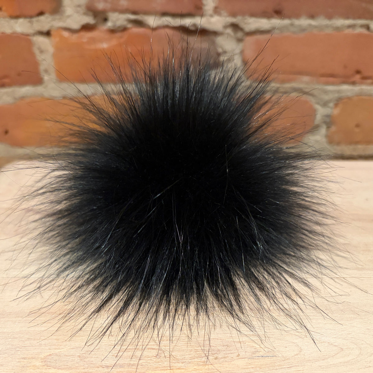 A large, fluffy black faux fur pom-pom resting on a light wood surface with a blurred red brick wall background.