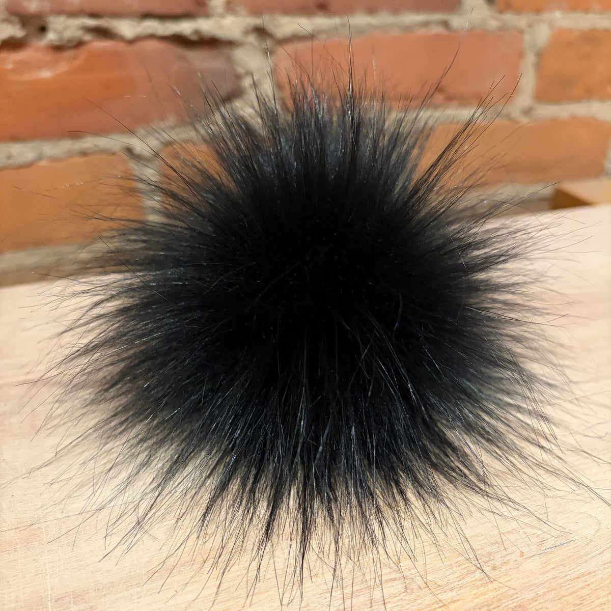 A black faux fur pom-pom ball resting on a light wood surface with a brick wall background.