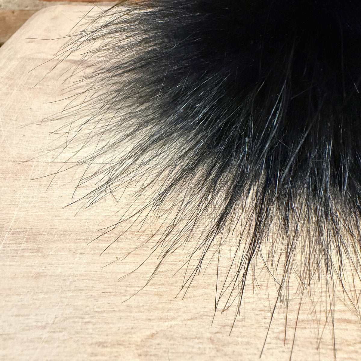 A close-up of a black fluffy faux fur trim or pom-pom resting on a light beige wood surface.