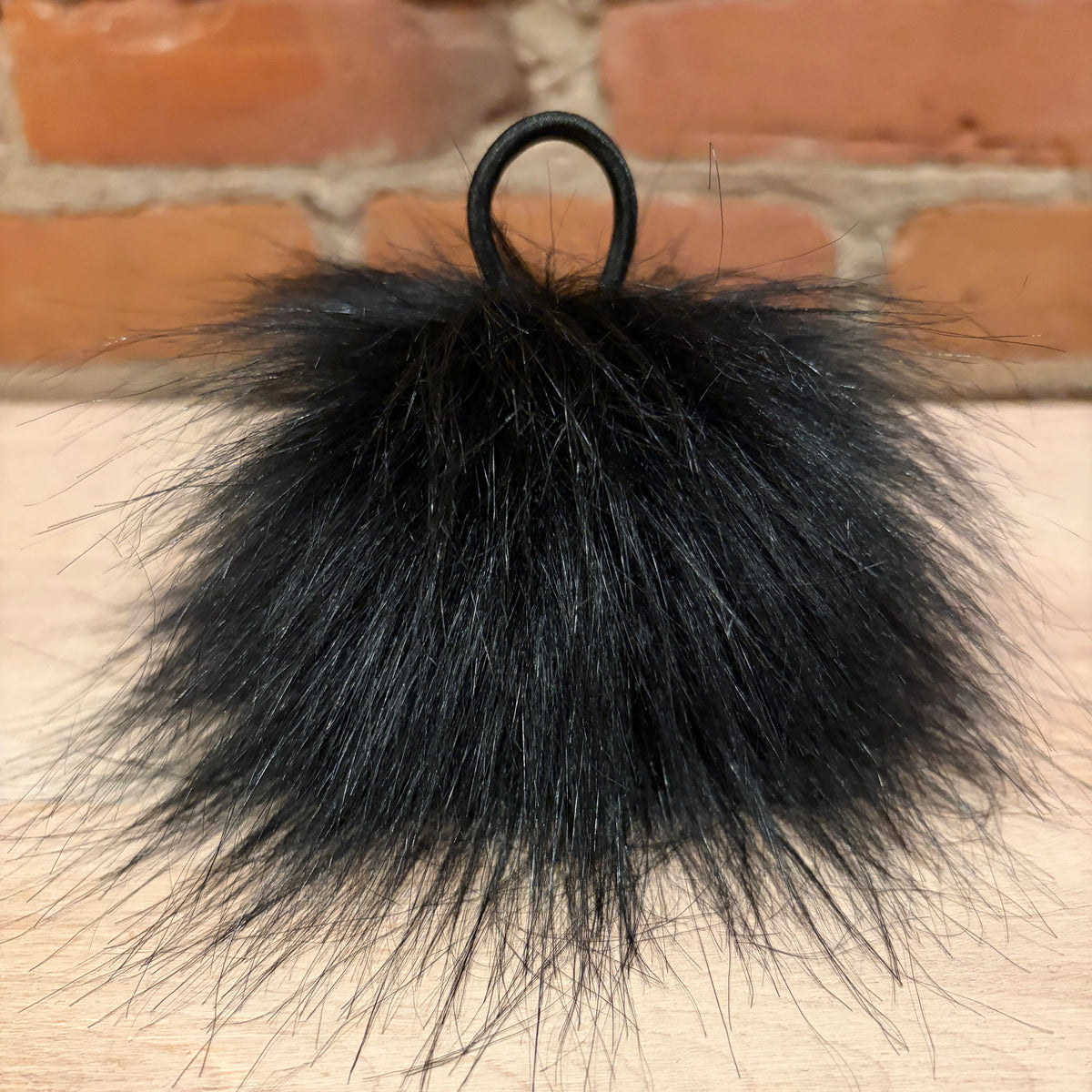 A fluffy black faux fur pom-pom keychain with a black elastic loop, resting on a light wood surface with a brick wall background.