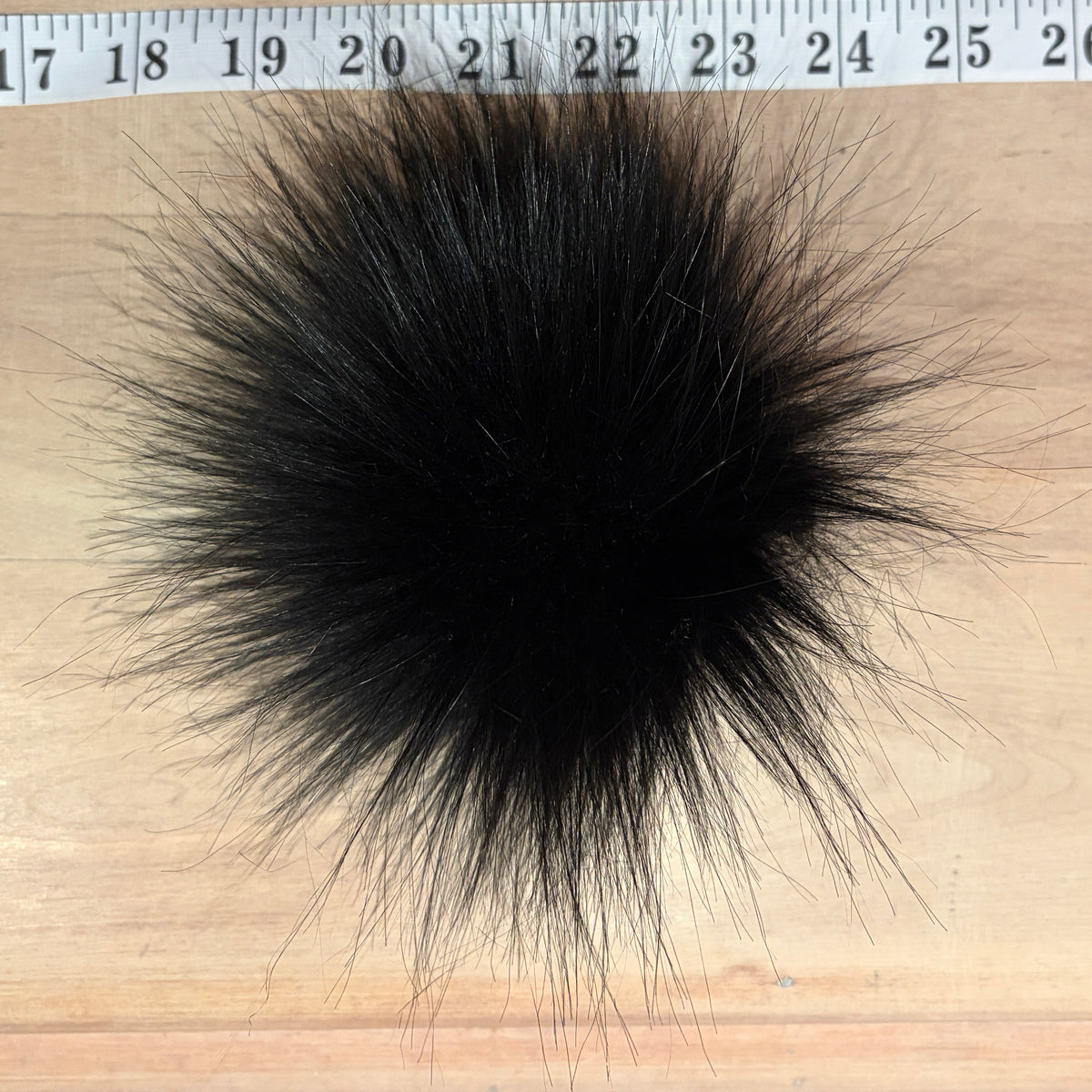 A black fluffy faux fur pom-pom or decorative ball placed on a light wood surface with a measuring tape visible along the top edge for scale.